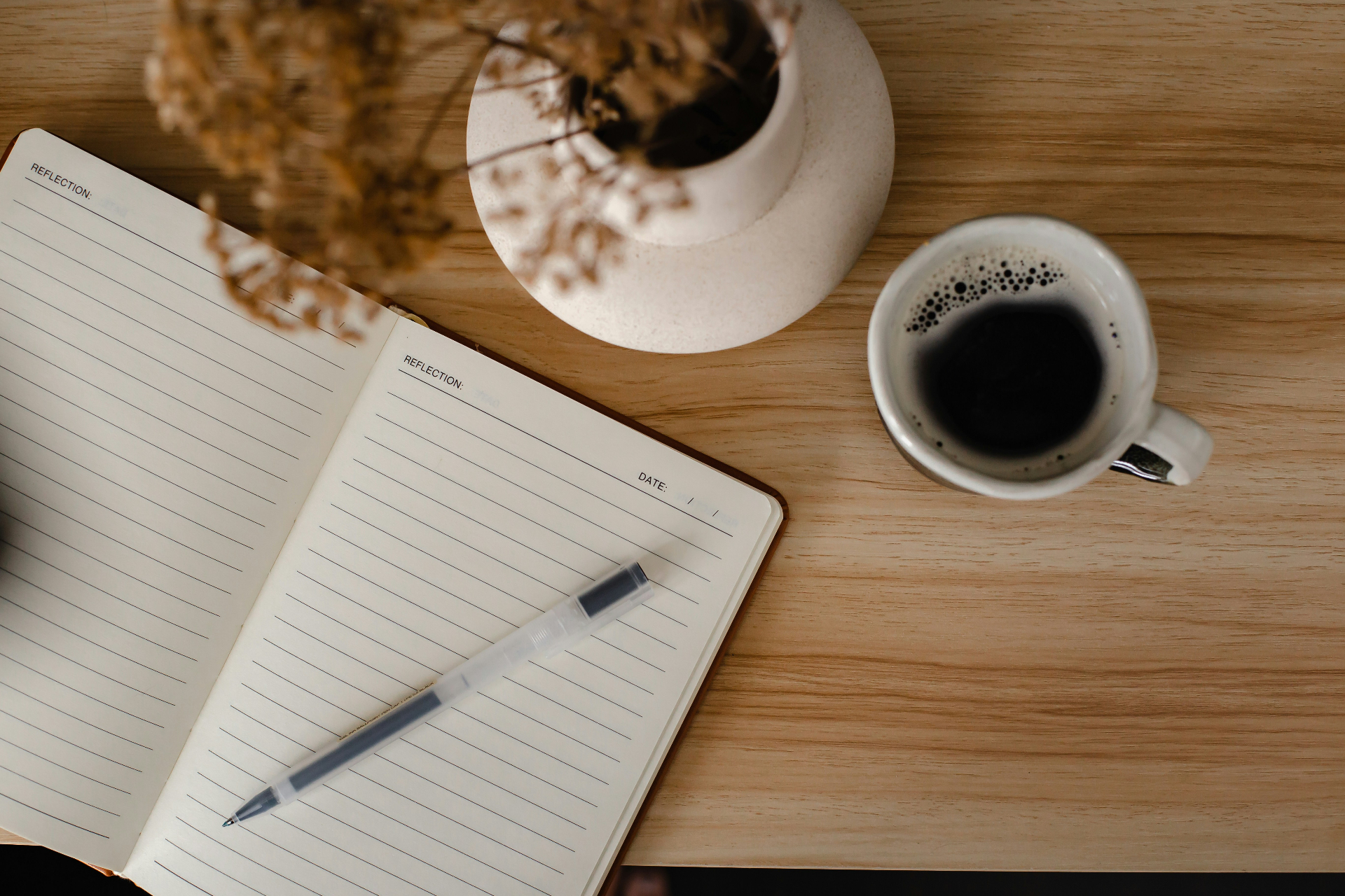 An open notebook with a pen beside a cup of coffee and a vase of dried grasses